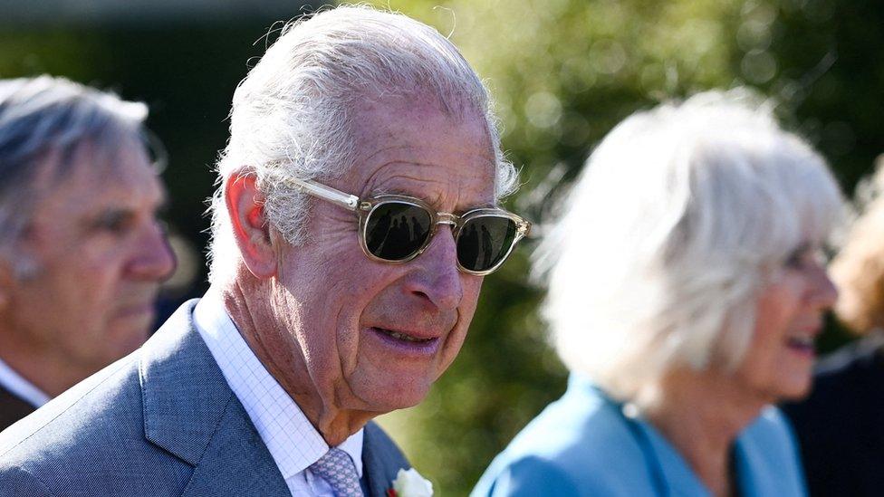 King Charles wearing sunglasses with Queen Camilla behind him