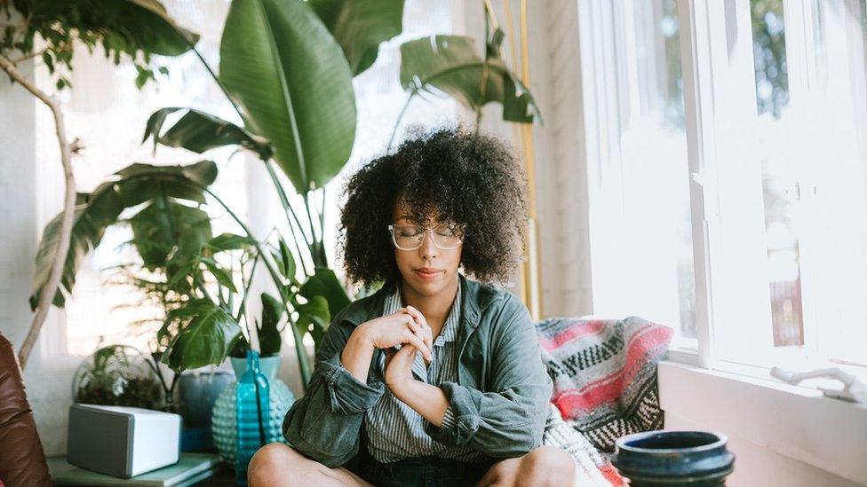 Young woman meditating on a sofa at home, plants on the background and a big, light, window to her left