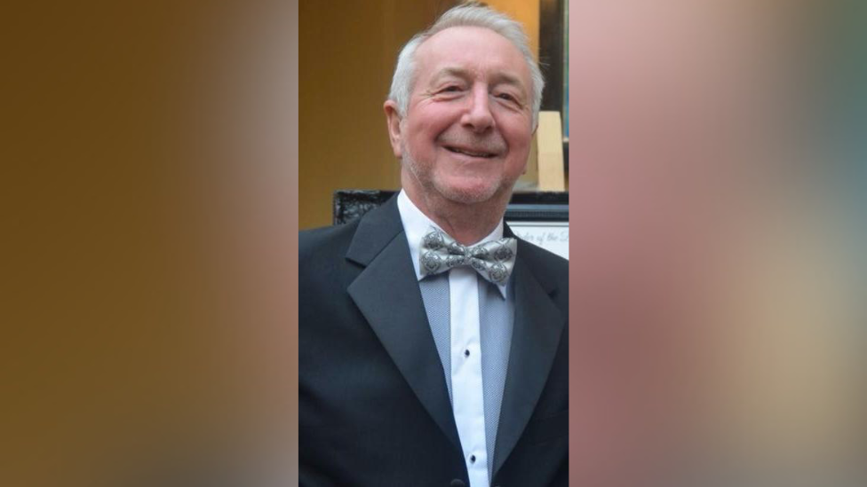 A man wearing a suit and a bow tie smiling into the camera. He is stood up.