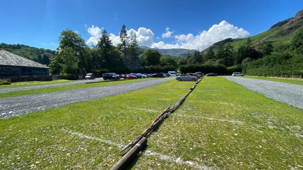 The car park in Coniston. White lines have been painted on grass while logs separate the bays.