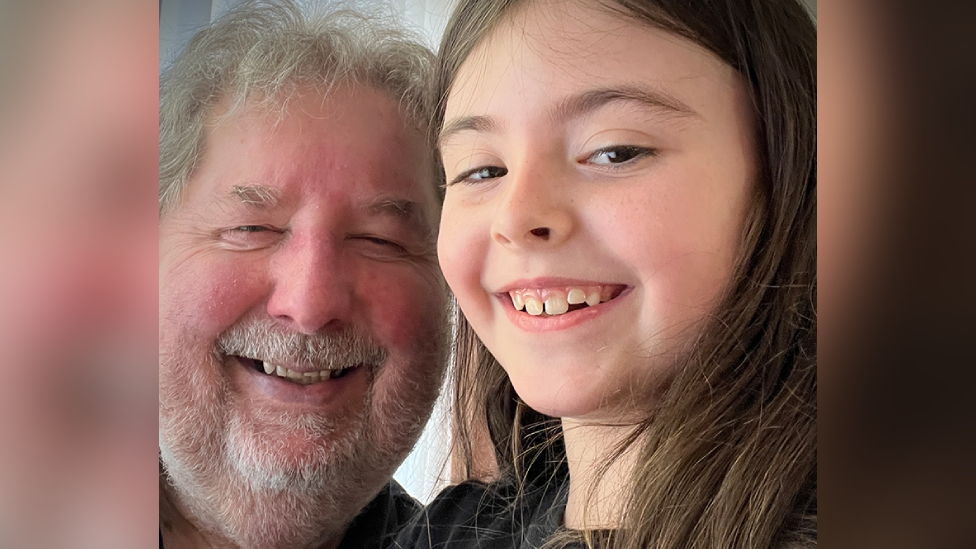A man with grey hair and beard smiling at the camera, next to him is a smiling ten-year-old girl with long dark hair