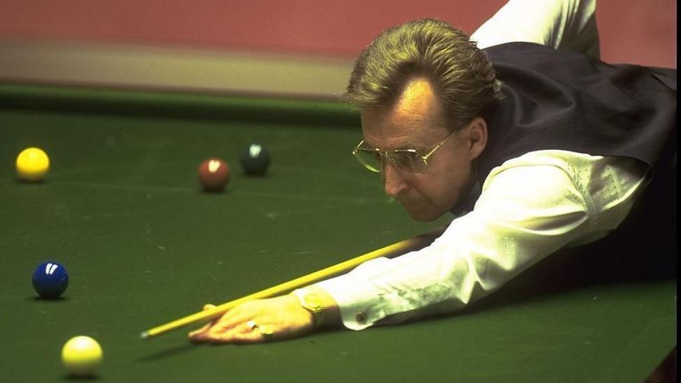 Terry Griffiths bent over the snooker table, cue resting on the bridge between his thumb and index finger on his right hand as he prepares to strike the white ball. He is wearing snooker attire, with a white long sleeve shirt and black vest. He also wears large and thick metal rim glasses, a gold wrist watch and has large rings on his ring and pinkie fingers 