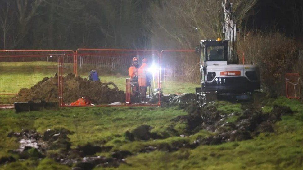 Muddy field with deep tracks left by white digger seen by metal fencing with two workers standing by a pile or earth. 