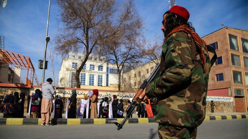 Taliban guard standing as women carry placards during a rally demanding the international community unfreeze the country's assets