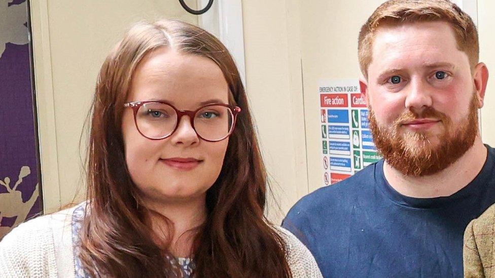 Tamar Vellacott and Cameron Pratt looking at the camera. Tamar wears glasses and has long hair, Cameron has ginger hair and a beard.