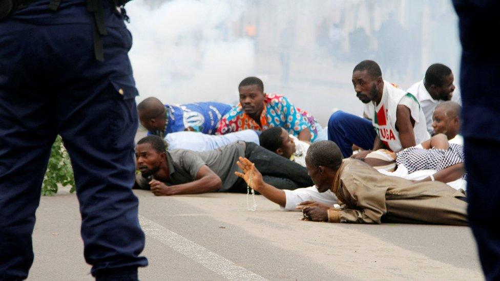 Protesters lie face down in front of security forces as clouds of tear gas swirl behind them. One man thrusts a hand forward, holding rosary beads aloft