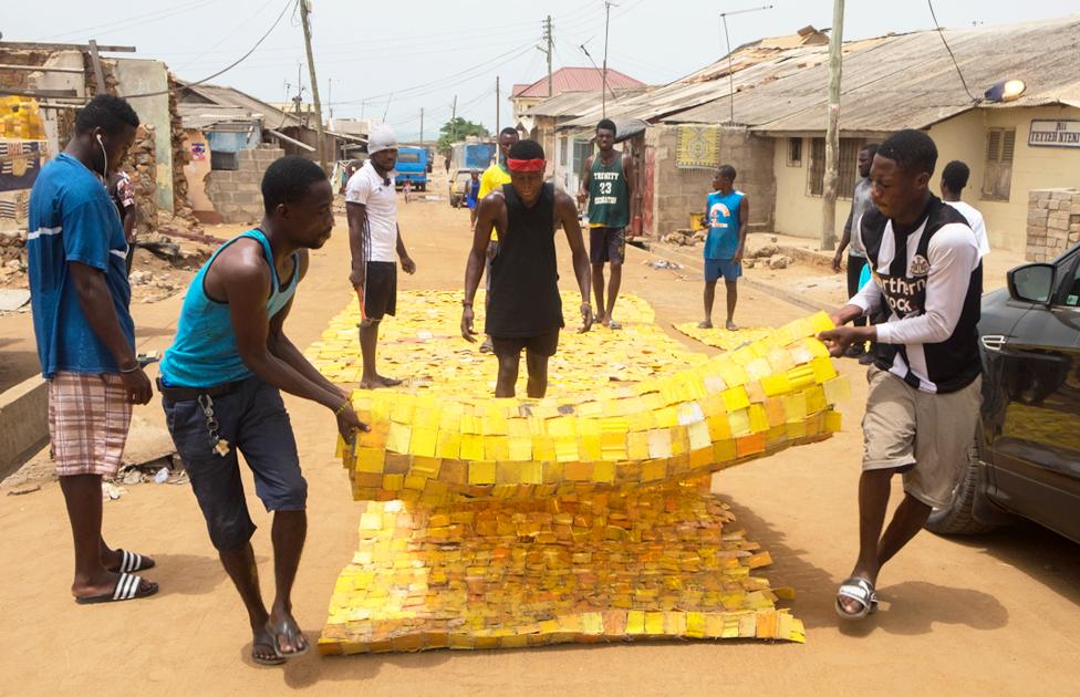 People rolling out yellow tapestry tapestry designed by artist Serge Attukwei Clottey on a road in La - Accra, Ghana