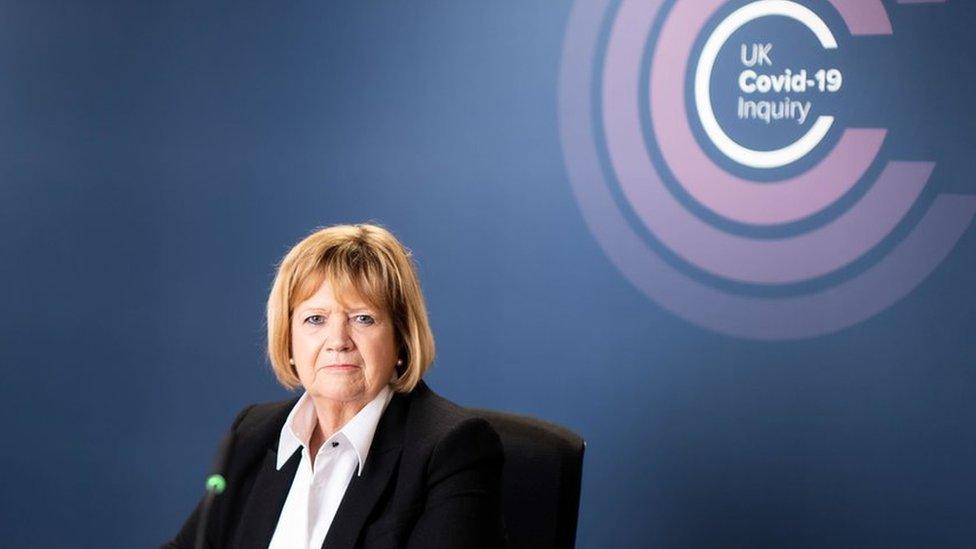 Baroness Heather Hallett white shirt and a back jacket while sitting at a desk in front of the UK Covid-19 inquiry logo, which is made up of several concentric pink circles.