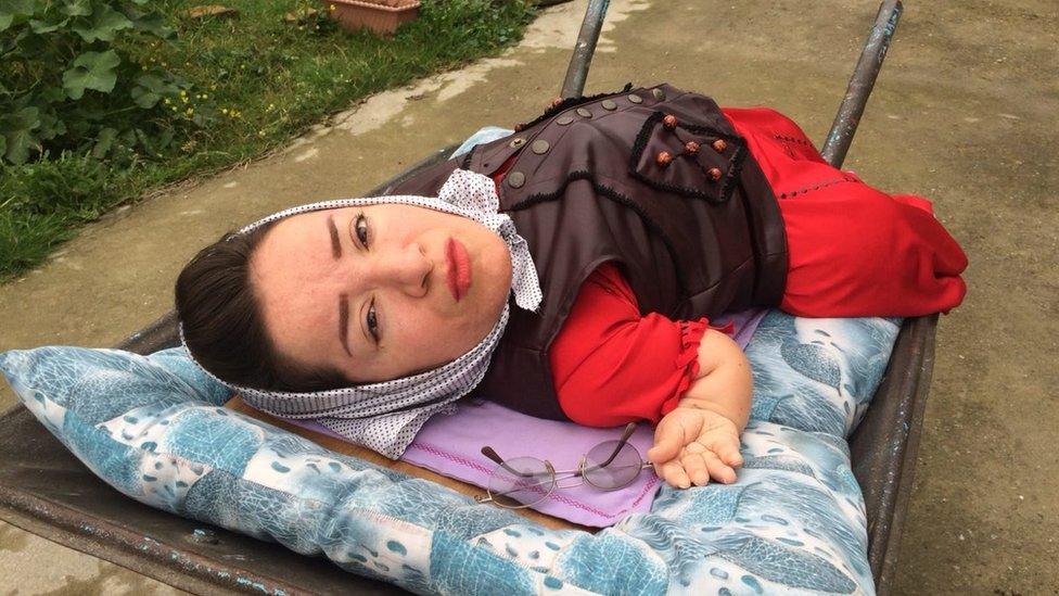 Mitra, a 41-year-old woman, living with 75 per cent physical disabilities on a cot. She is wearing a red dress and a brown jacket in the picture.