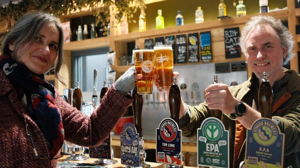 Jojo Mehta and Greg Pilley standing at a bar holding pints in the air