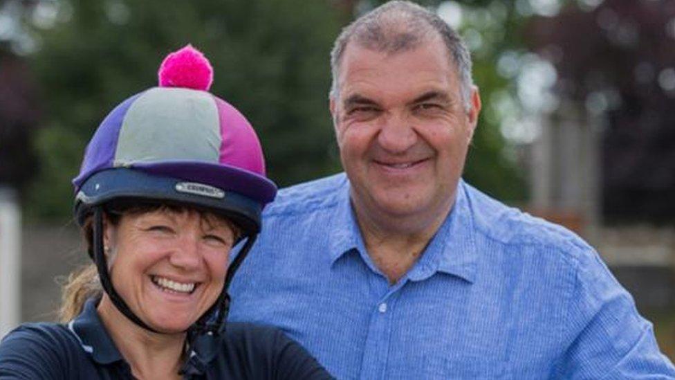 A woman in a horse riding hat and a man with grey hair both smiling at the camera