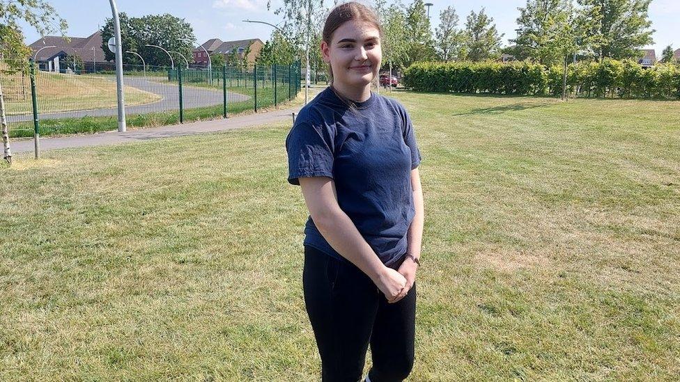 Fay, standing in a park, she is slightly smiling at the camera, she is wearing a blue top and black leggings.