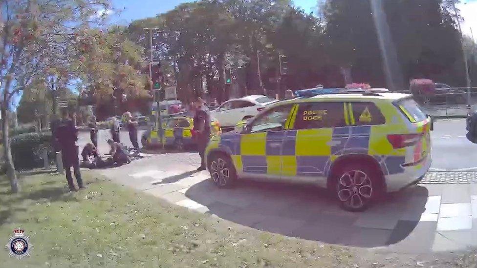 Police car alongside a group of police officers who are arresting an offender who is lying on the ground