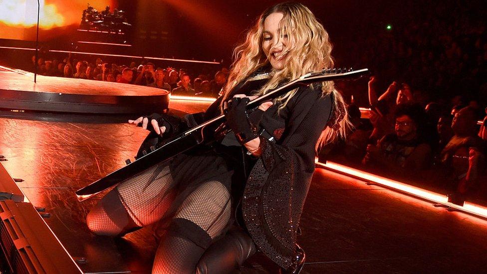 Singer Madonna playing guitar and smiling while on her knees on a stage