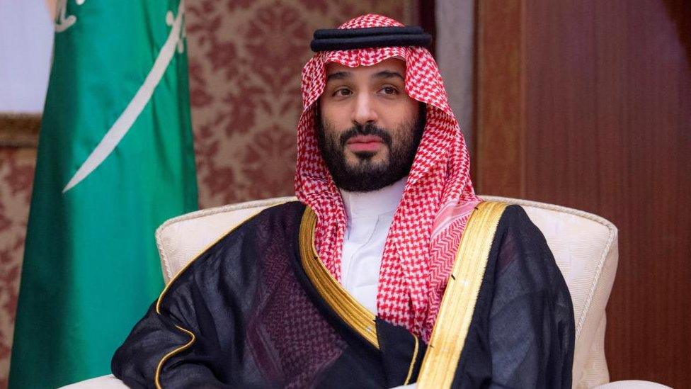 Saudi Arabia's Crown Prince Mohammed bin Salman sits in a large white chair at a summit. A Saudi flag can be seen hanging in the background.