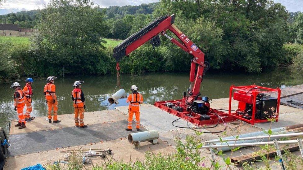 Several men in orange work outfits next to a small crane, standing on a pontoon on the river