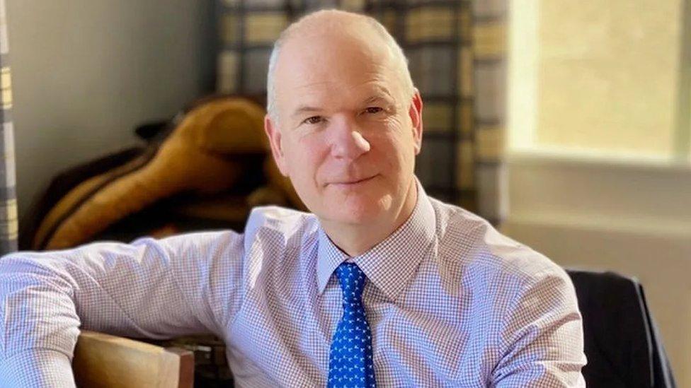 Mark Shelford in a checked shirt and blue tie, sitting on an armchair