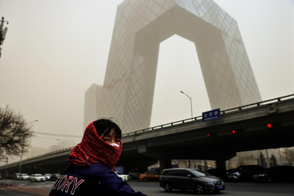 A woman wearing a head covering is seen in front of the headquarters of China's state media broadcaster CCTV that is shrouded in haze after a sandstorm in the Central Business District of Beijing, on 15 March 2021