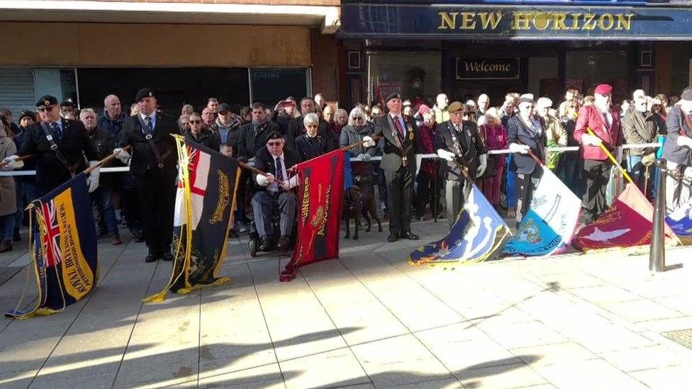 Flags in various colours are lowered by uniformed representatives, with a crowd of people behind them