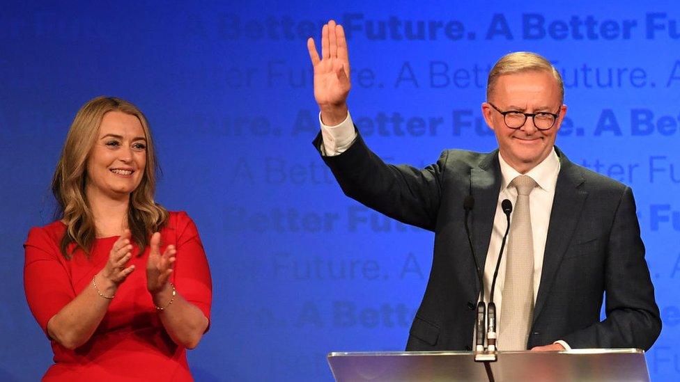 Anthony Albanese waves from behind a lectern beside his partner