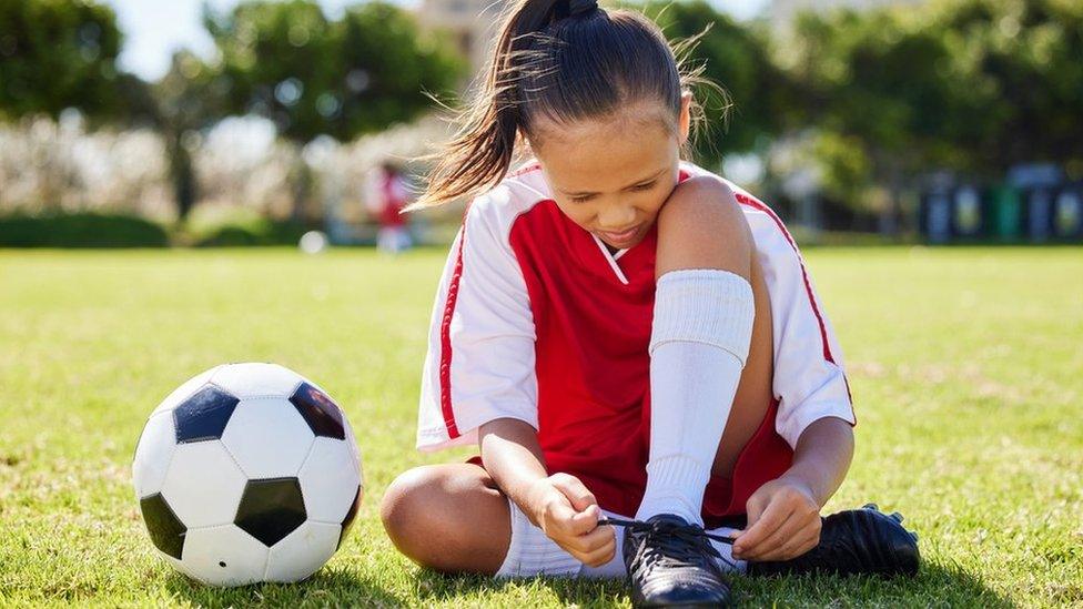 Girl ties football shoes sat down with ball next to her in the park