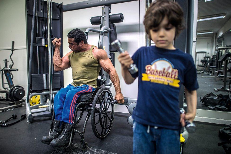 A man lifts weights in a gym while a small child lifts another one.