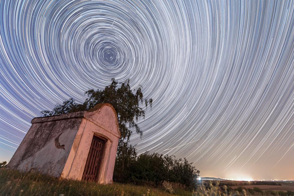 A combination of multiple long exposure photographs shows satellites and Perseid meteors crossing a starry sky over a chapel