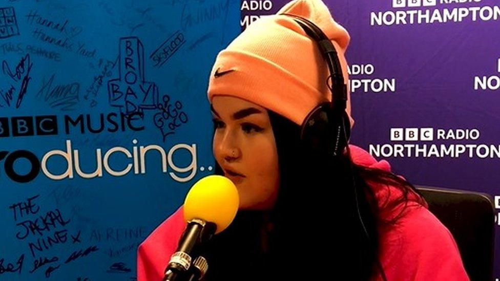 Mae Stephens wearing an orange hat, a pink top and headphones talking into a microphone with BBC Radio Northampton branding in the background