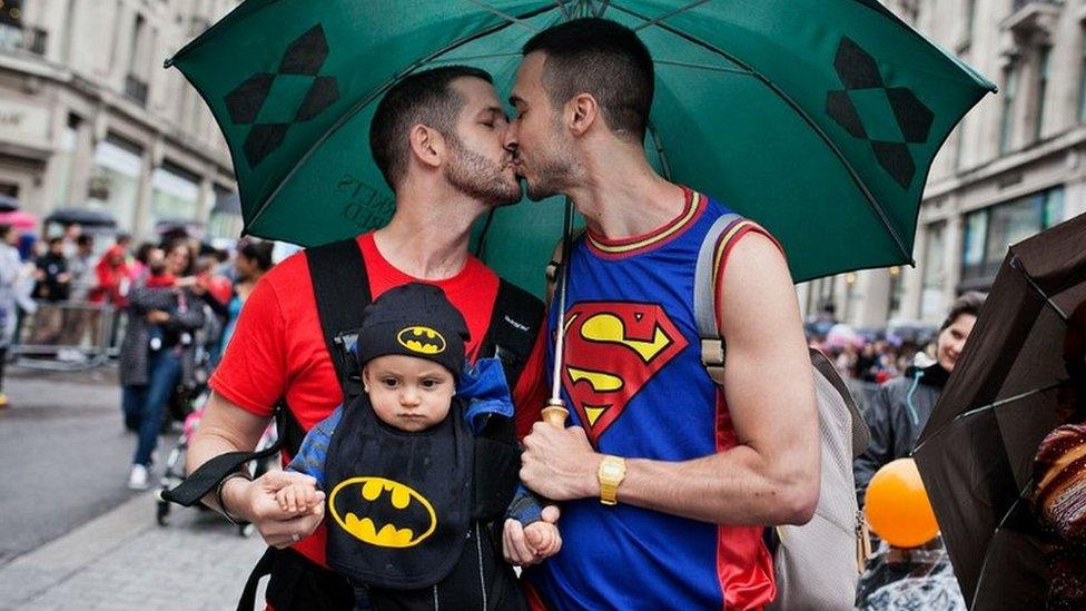 Same-sex couple kiss with one of them holding a baby on his chest