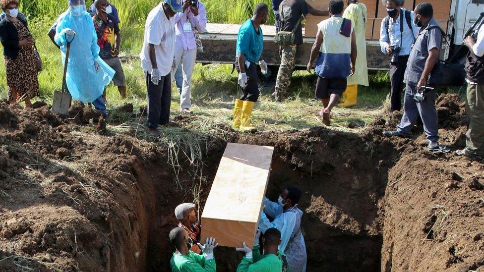 Workers wearing personal protective equipment put a casket into a deep grave