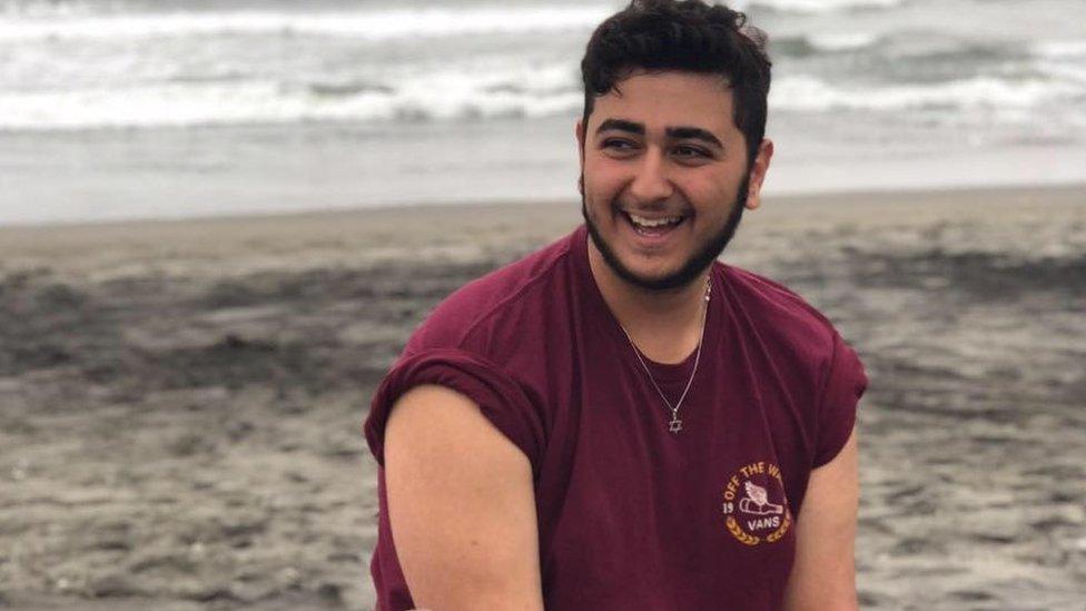 Young man with beard and red shirt sitting at the shore, water in background