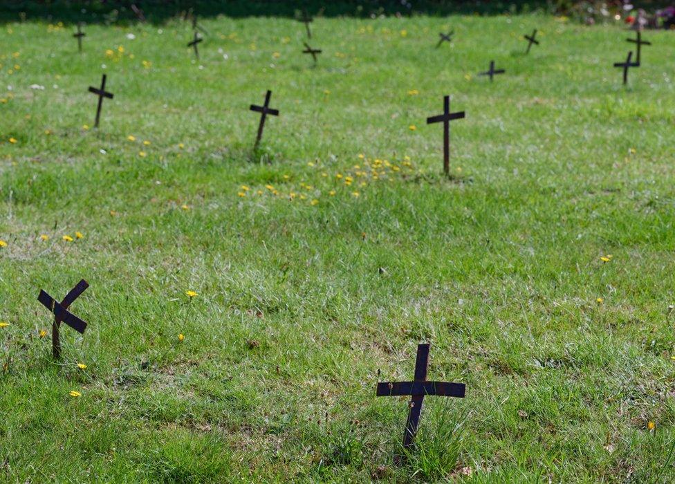 A large number of small iron crosses across the grass of Nayland cemetary. A few dandelions are growing up around the crosses and in the distance, to the top right of the picture, are floral tributes left in front of standard gravestone