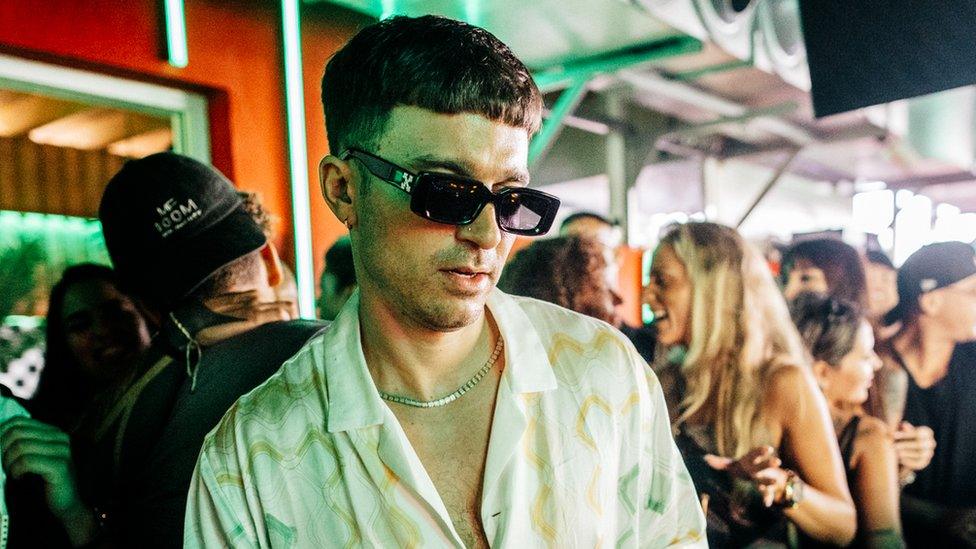 Michael Bibi DJing with people dancing in the background. He is wearing a white shirt and dark sunglasses and is looking down at the DJ decks