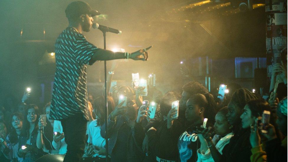 6lack stands in front of a crowd of young apprentices who are recording the show on their phones