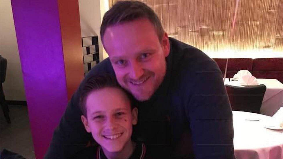 A father and son in a restaurant. The father stands over his son with his arms round him.