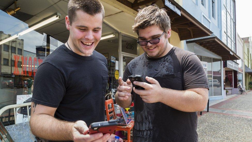 two men standing next to each other and playing on their phones