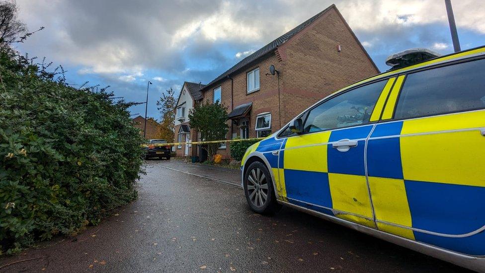 A police car in the foreground with a cordoned-off house in the background