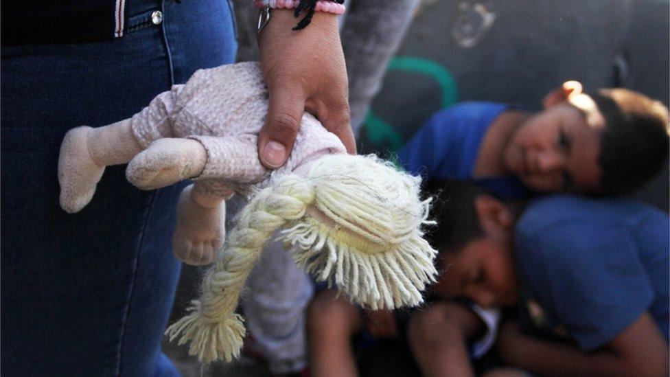 Woman holds doll at US-Mexico border, children resting behind her