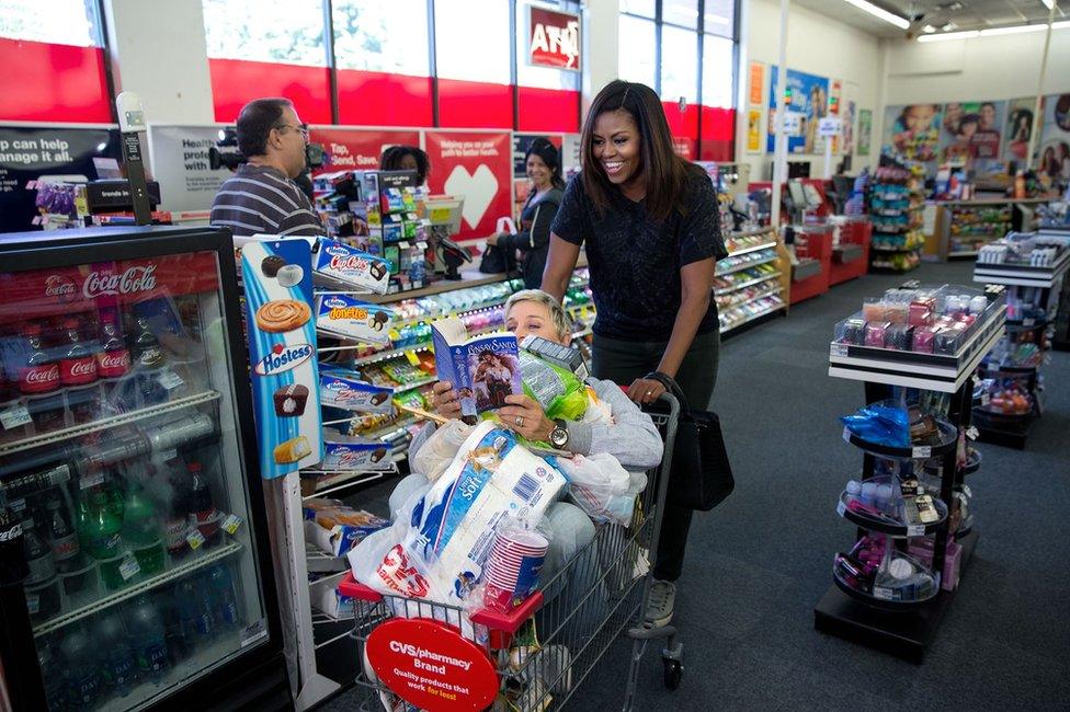 Michelle Obama wheels a loaded trolley - also containing Ellen de Generes - through a pharmacy. Ms De Generes is reading a romance novel while sitting under a selection of groceries.