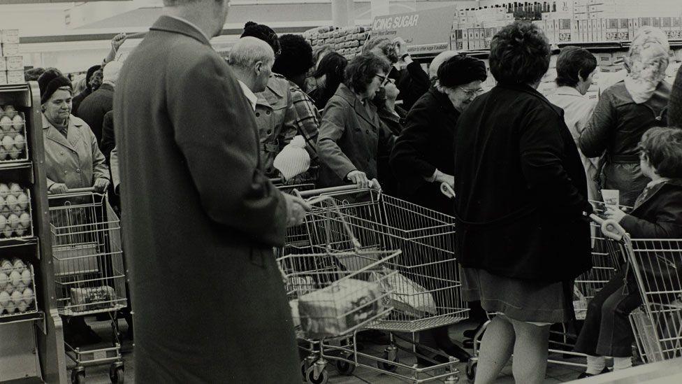 A black and white image of men and women in 1974 in a supermarket, pushing supermarket trolleys and looking at a just seen pile of sugar in front of them. Above it is a sign saying icing sugar