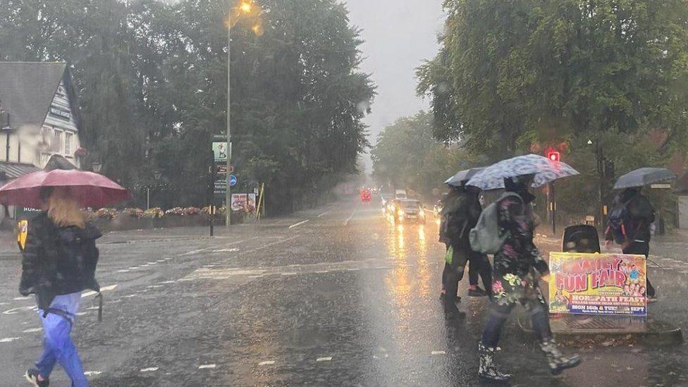 Rain-soaked road with people in wellingtons with umbrellas crossing