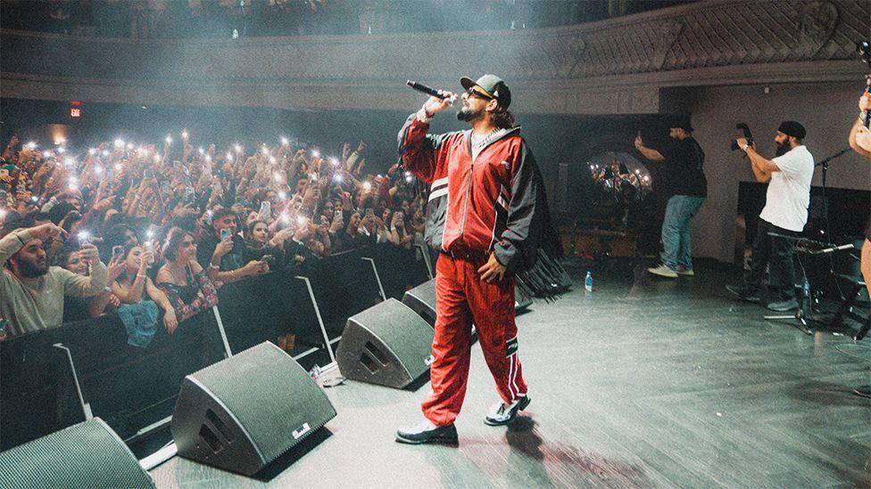 Raf, dressed in a red and black tracksuit-top combination, sunglasses and black hat, singing into a microphone to fans who have their phones up with the torch light on. Raf is standing on a stage, which has black speakers at the front, with members of the crew behind him taking photos.