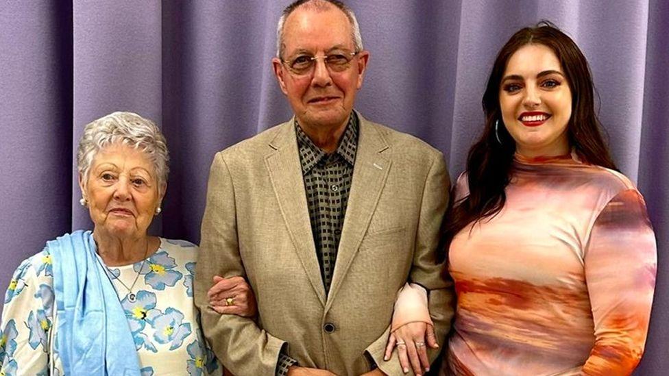 Marilyn, who has short white hair and is wearing a blue floral dress, is on the arm of Ron who is smiling in a beige suit. He is wearing glasses and he has short grey hair. Next to the couple is Olivia who has long, dark hair. She is arm-in-arm with her grandfather. All three are standing in front of a light purple curtain.