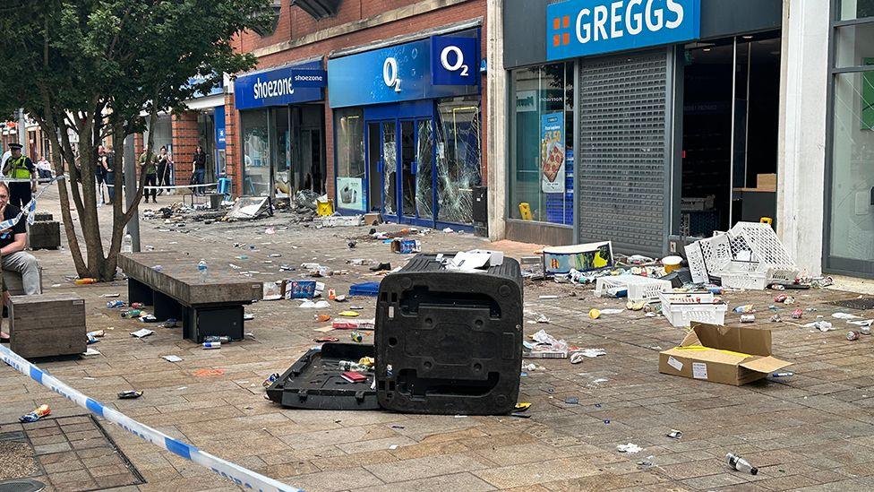 Rubbish lies outside of a row of shops with damaged windows