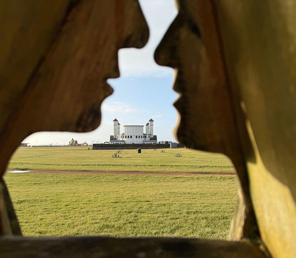 A view between two statues which look as if they are about to kiss reveals a broad expanse of grass and, in the distance a white building with several towers against a blue sky