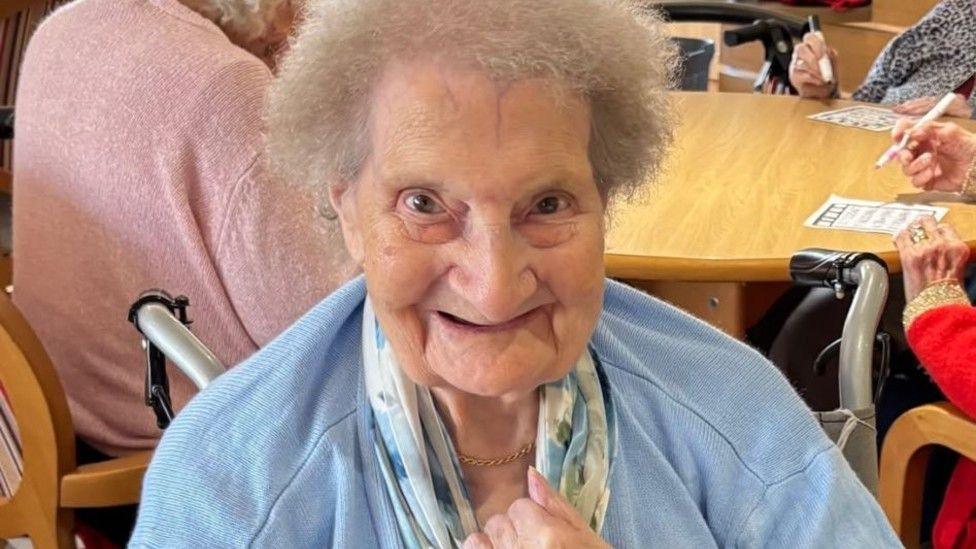 A close-up of an elderly woman smiling at the camera. She is wearing a light blue cardigan and a scarf.