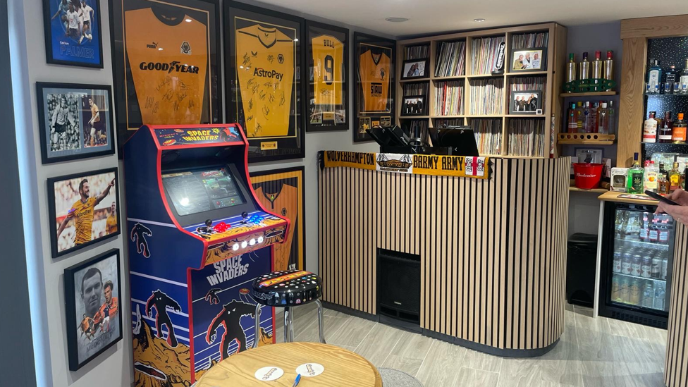 Wolves memorabilia fills the walls of the pub alongside a games machine and stools and tables. A small fridge is filled with drinks on the right side and spirits are on the wall, with an ice bucket. A DJ booth is on the left of the time with wooden panelling on the front and shelves filled with record behind.