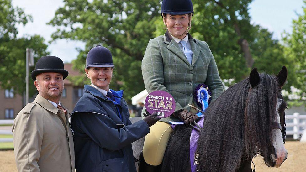 Beth Jones sitting on Blaze, a pony with a long black main and white blaze down his nose. She is wearing jodhpurs, a navy riding hat and a green checked riding jacket. To the left a man and woman dressed in smart coats and hats are handing her a round purple plaque that says "Search 4 a star"