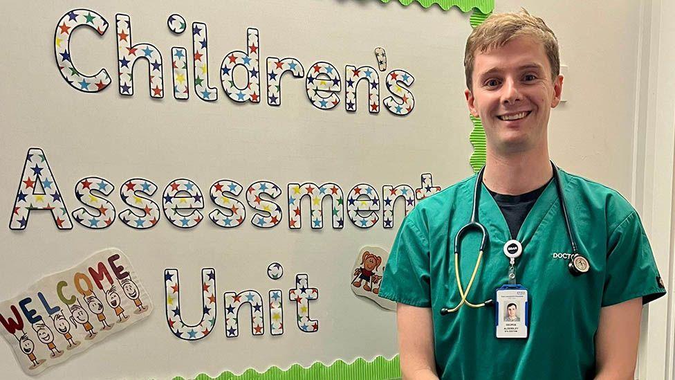 Dr George Aldersley, in green scrubs and with a stethoscope round his neck, standing smiling in front of a sign that reads Children's assessment Unit