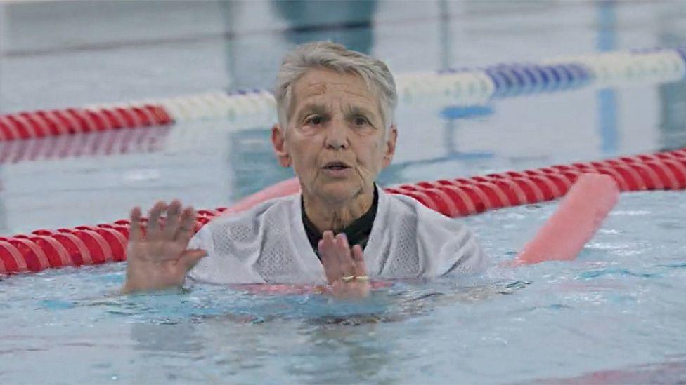 Alison Hubbard in a swimming pool, with a white T-shirt on and floating with the help of a red cylindrical buoyancy aid. She is gesturing to other people in the pool (out of shot), with her hands slightly raised.